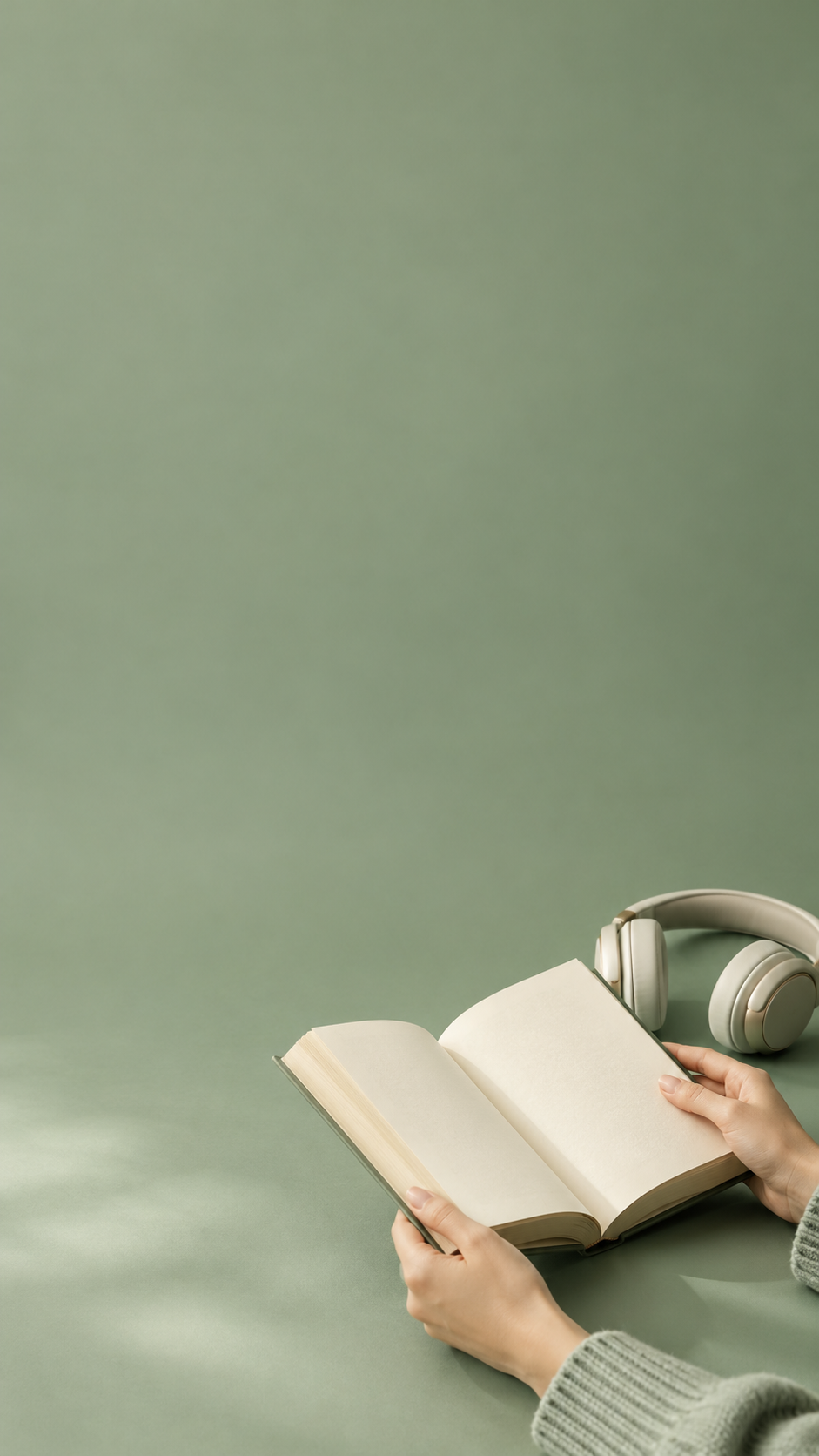 Hands holding an open book beside headphones on a sage green surface